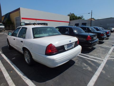 2003 Ford Crown Victoria - Rear three-quarter view of a white Ford full-size sedan parked beside a line of black sedans in a paved lot by a red-striped building.