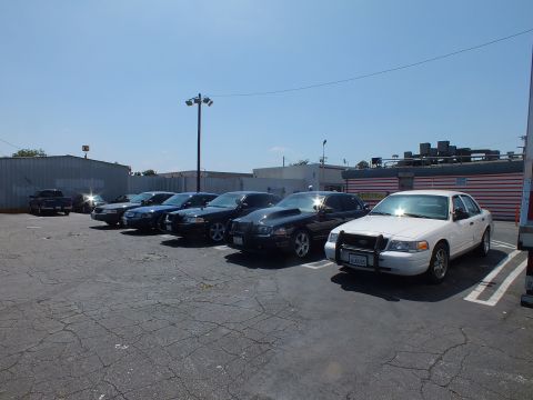 2003 Ford Crown Victoria - row of full-size sedans, including a white Ford with a police-style push bumper, parked across a cracked asphalt lot.