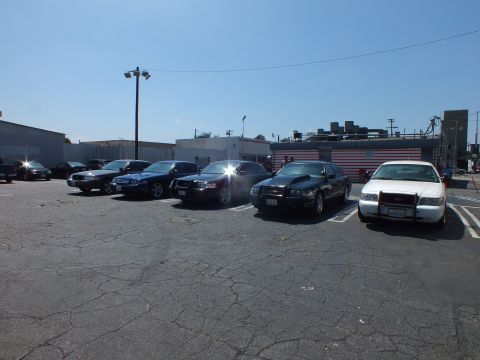 2003 Ford Crown Victoria - wide view of several dark sedans and a white car parked in marked spaces across a sunlit asphalt lot.