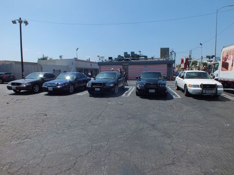 2003 Ford Crown Victoria - Five full-size sedans, mostly dark with one white, line up facing the camera in a parking lot beside a striped building.
