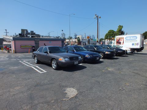 2003 Ford Crown Victoria - gray Ford sedan leads a row of parked cars near a striped building and a Hill-Rom box truck in an urban lot.