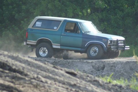 1996 Ford Bronco - Side view of a teal and blue two-tone square-body Ford Bronco with a tan removable hardtop running through a dirt pit, throwing dust off th…