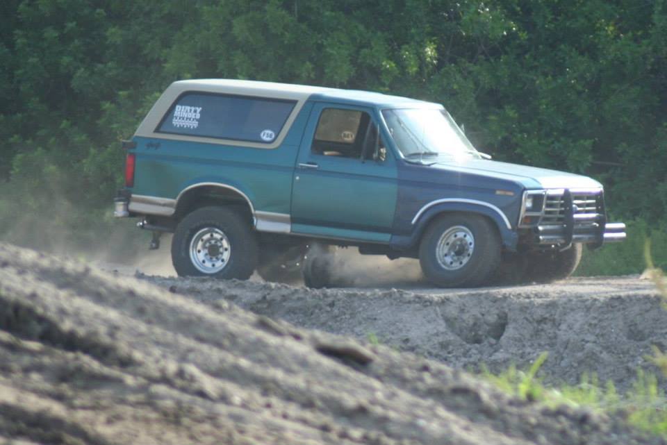 Teal square-body Ford Bronco with tan top kicking up dust in a dirt pit, side view