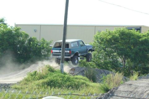 1996 Ford Bronco - A teal Ford Bronco is captured mid-air during a jump on a dirt track, kicking up dust.