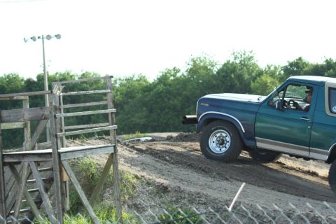 1996 Ford Bronco - A teal Ford Bronco navigates a dirt track, with a wooden ramp structure visible to the left.
