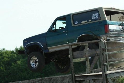 1996 Ford Bronco - Teal and tan two-tone full-size Ford Bronco with removable hardtop climbing a wooden ramp obstacle, front wheels airborne against a bright…
