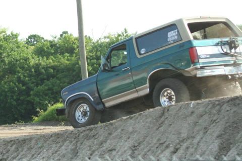 1996 Ford Bronco - Teal-green two-tone square-body Ford Bronco with a tan removable hardtop dropping nose-down off a steep dirt bank, rear wheels still on the…