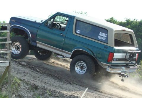 1996 Ford Bronco - A lifted teal-green 1990s Ford Bronco with a white roof climbs a dirt berm at an angle, front left wheel lifted and dust kicking up behind…