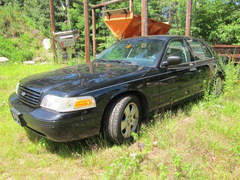 2001 Ford Crown Victoria - A black 2001 Ford Crown Victoria sedan is parked in a grassy, outdoor area with some industrial equipment in the background.