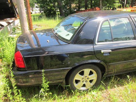 2001 Ford Crown Victoria - The rear of a black 2001 Ford Crown Victoria sedan is visible, parked in tall grass with a wooden structure to its left.