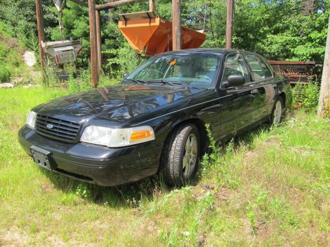 2001 Ford Crown Victoria - A black sedan is parked in a grassy area with some industrial equipment in the background.