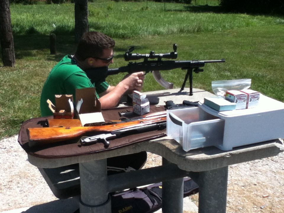Man in green shirt aims rifle with scope at outdoor range, table with ammunition and gun case
