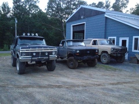 Three Ford trucks parked in front of a blue building. The truck on the left appears to be a 1979 Ford Bronco,