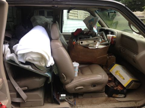 Interior of a tan SUV filled with boxes, blankets, and car parts. A cup sits in the center console