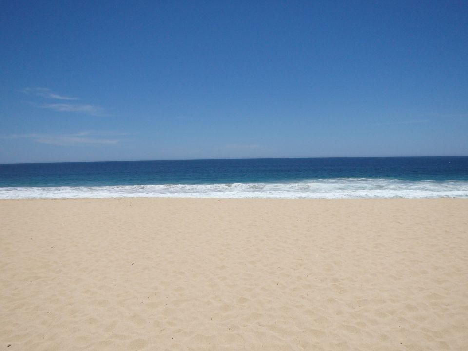Sandy beach meets blue ocean under a clear blue sky. Water has white waves