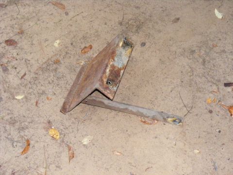 1978 Ford Bronco - A single, heavily rusted metal bracket lies on a dirt surface with scattered leaves.