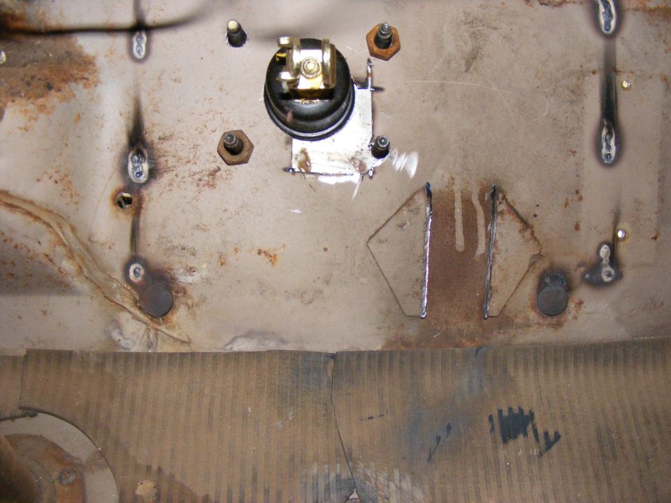 Close-up of a vehicle's firewall with a black electrical component mounted on a metal plate, surrounded by bolts and welds
