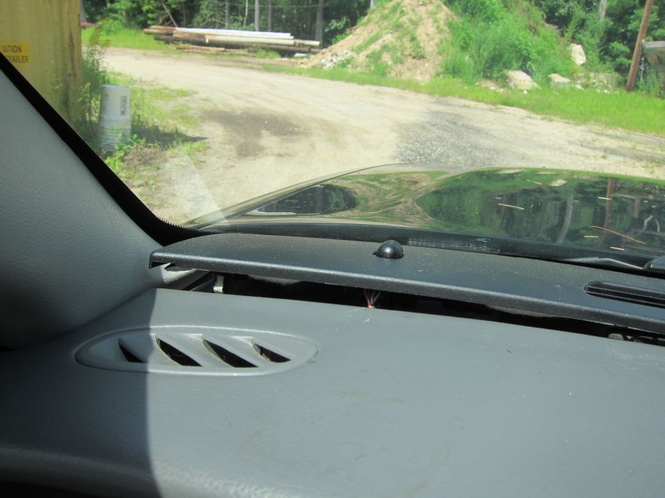 Dashboard interior showing air vents and part of the windshield. A dirt road and trees are visible outside