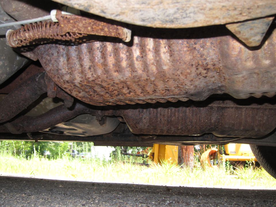 Underside view of a vehicle showing a rusty catalytic converter and exhaust pipes. A spring mechanism is visible near the top