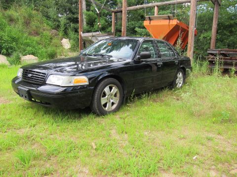 2001 Ford Crown Victoria - A black 2001 Ford Crown Victoria is parked in a grassy area with trees and industrial equipment in the background.