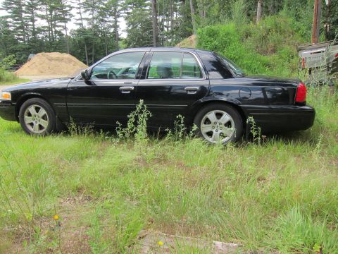 2001 Ford Crown Victoria - A black 2001 Ford Crown Victoria sits parked in an overgrown grassy area with trees in the background.