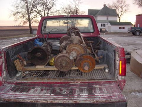 1992 Ford Bronco - A red pickup truck carries a load of assorted used car parts, including axles and brake drums.