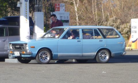 1969 Lincoln Continental - A light blue station wagon is parked at a gas pump, with people inside and a man standing nearby.