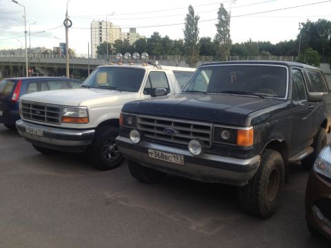 1987 Ford Bronco - Two Ford Broncos, one dark blue and one white, are parked on a street.