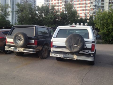 1987 Ford Bronco - Two Ford Broncos are parked in a lot, with apartment buildings in the background.
