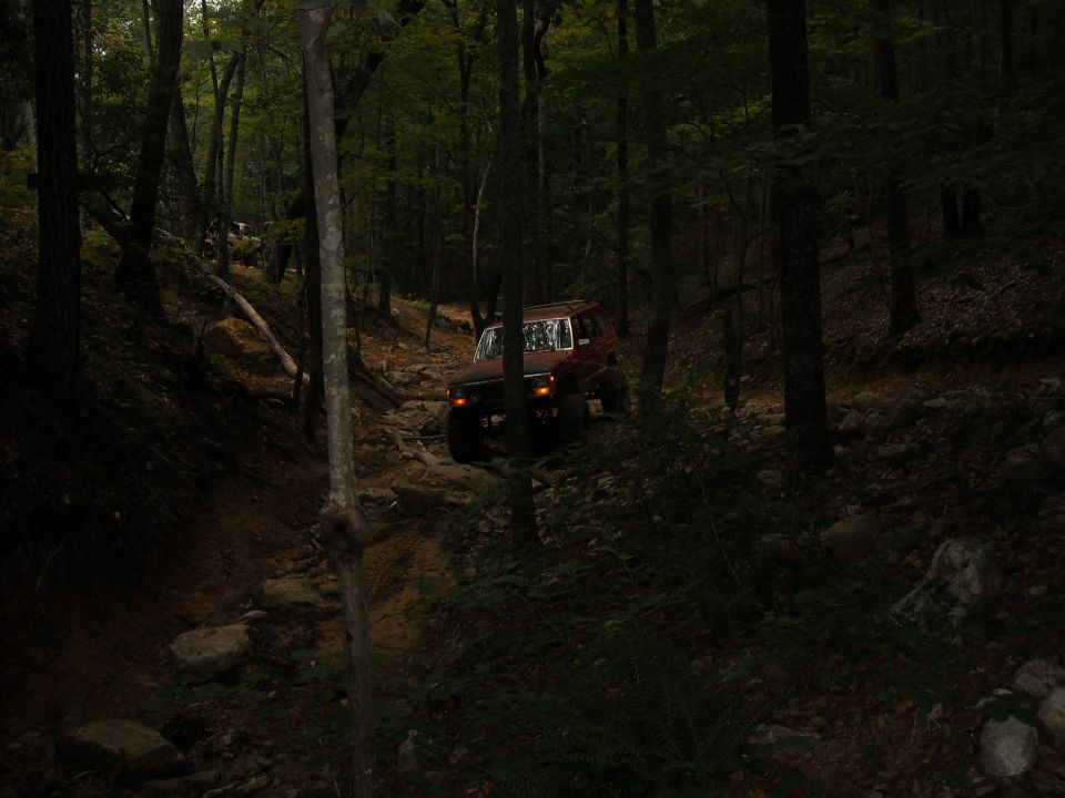Red off-road vehicle navigating a steep, rocky trail through a dense forest