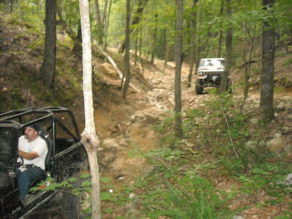 Two off-road vehicles on a rocky, wooded trail. The foreground vehicle is black with a roll cage and a driver. The background vehicle is white