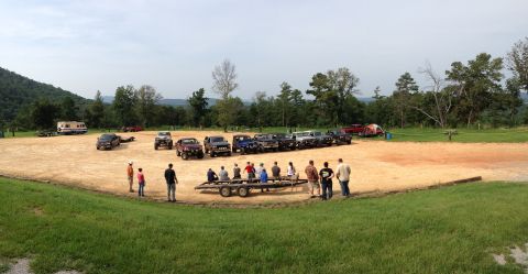 1989 Ford Bronco - A group of people stand around a trailer in a dirt lot with multiple SUVs and trucks parked nearby.