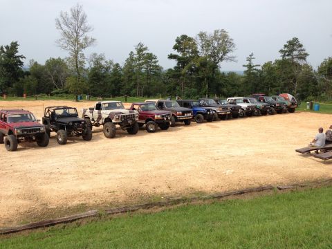 1989 Ford Bronco - A row of various off-road vehicles are parked on a dirt lot with trees in the background.
