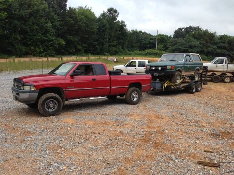 1989 Ford Bronco - Red Dodge Ram pickup tows a green Ford Bronco on a trailer, with another white pickup in the background.