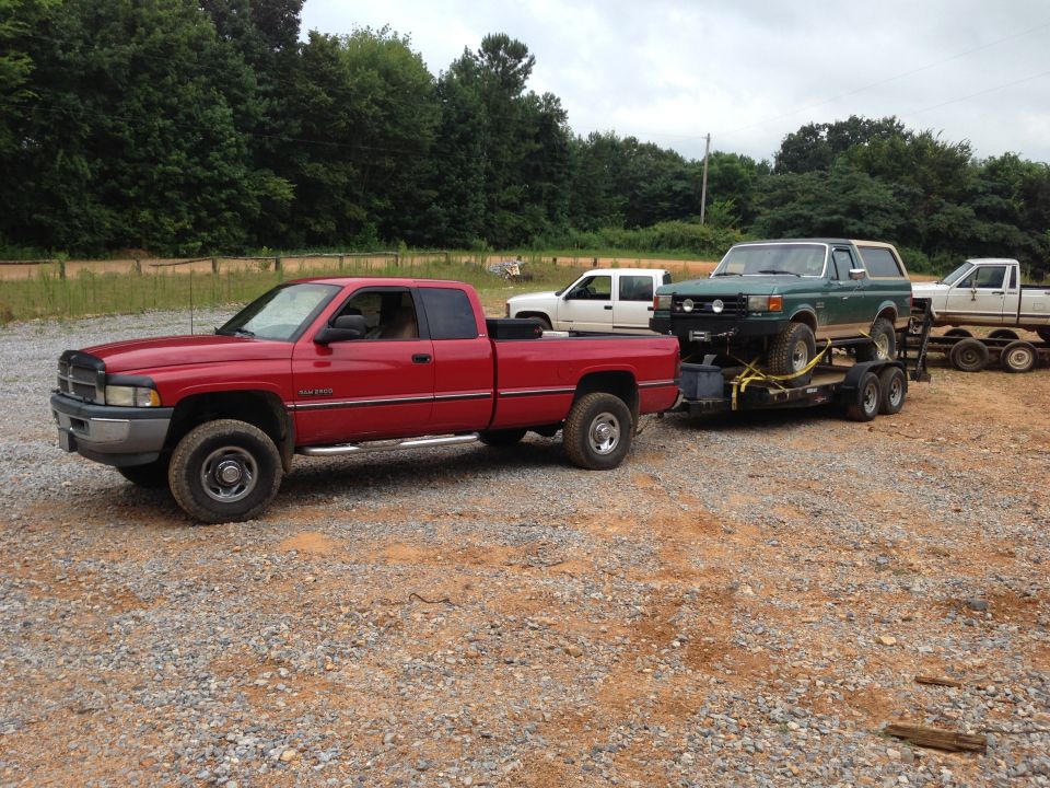 Red Dodge Ram pickup truck towing a green Ford Bronco on a trailer; white pickup truck in background