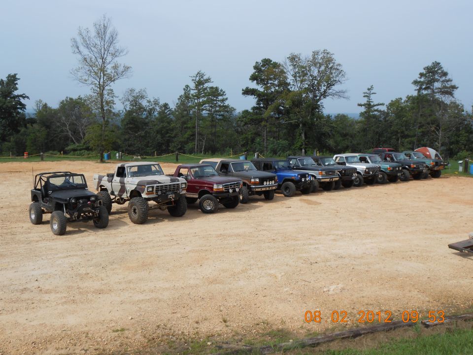 Line of modified off-road trucks and SUVs parked on dirt lot with trees in background