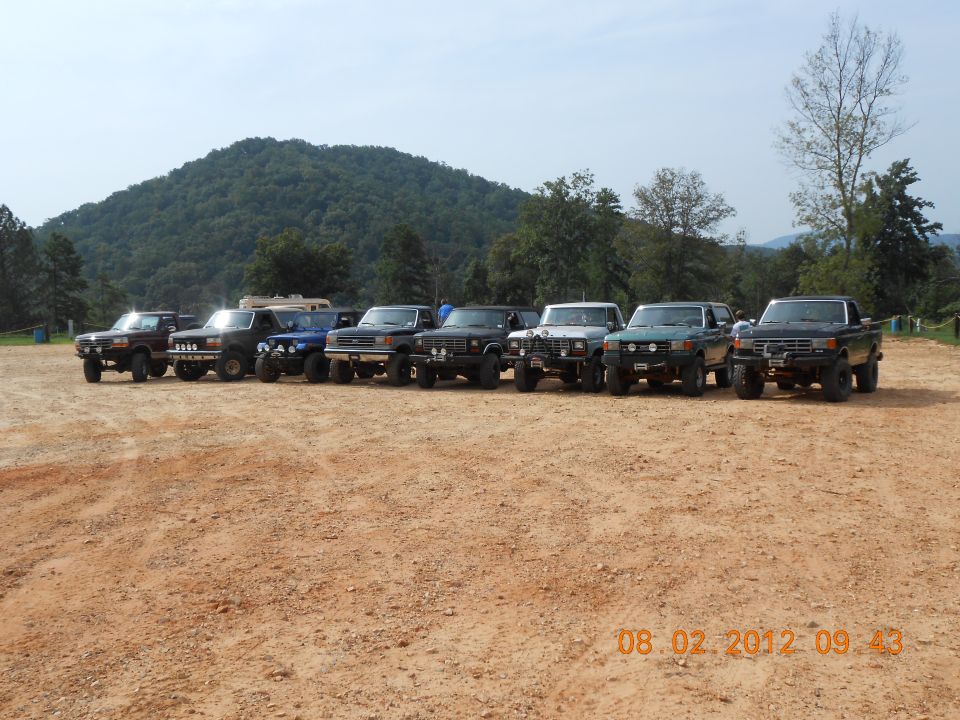 Line of modified off-road trucks and SUVs, mostly from the 1980s, parked on a dirt lot with a hill in the background
