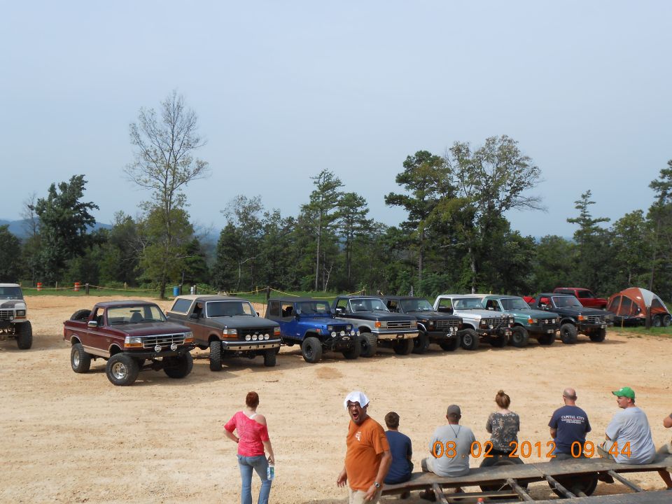 Several lifted trucks and SUVs parked on dirt lot with trees in background. One maroon truck in foreground