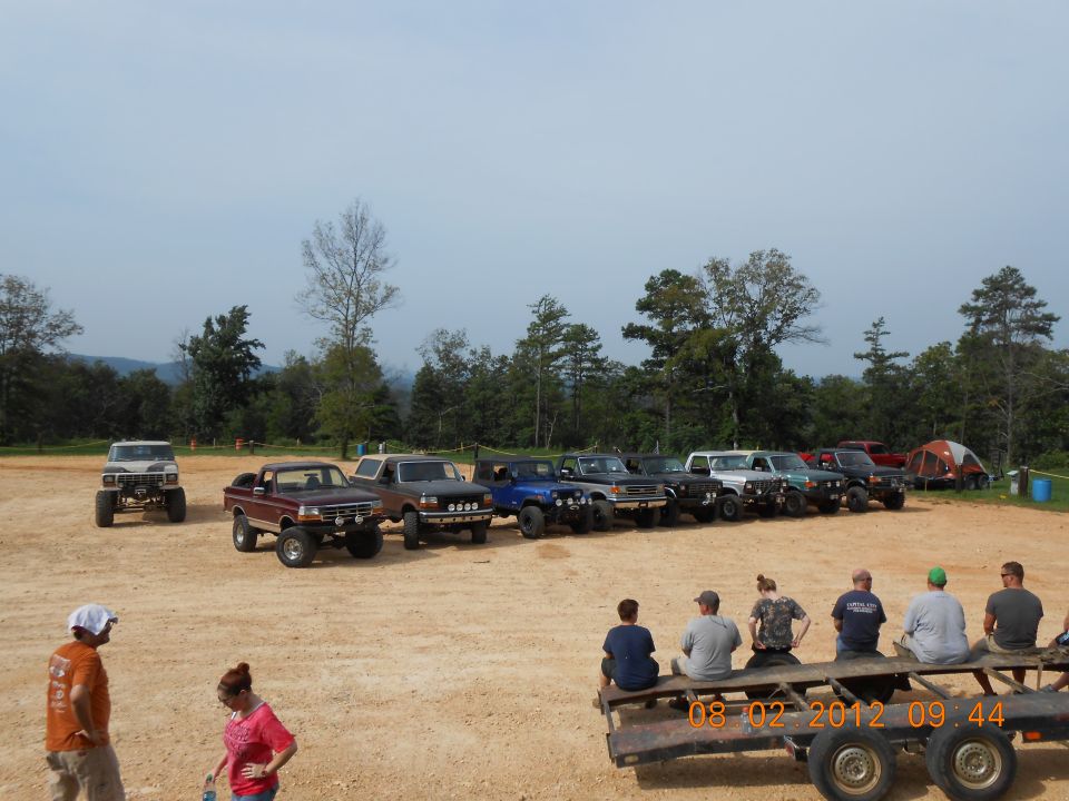 Several off-road trucks and SUVs are parked in a dirt lot with trees and hills in the background. Some people are sitting on a trailer