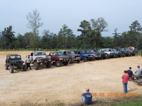 2013 Ford Bronco - A collection of modified 1980s Ford trucks and SUVs are lined up on a dusty lot with trees in the background.