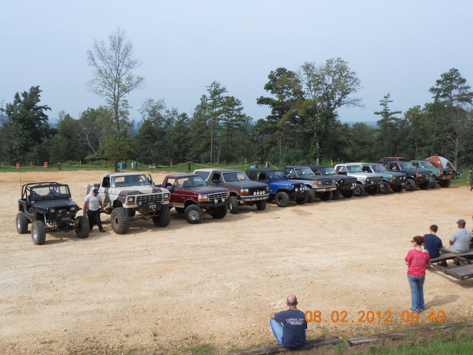 Line of lifted off-road trucks and SUVs, mostly Ford Broncos and F-Series trucks from the 1980s, parked on a dirt lot