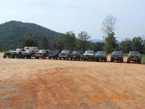 2013 Ford Bronco - Several modified off-road vehicles are parked in a line on a dirt lot with a wooded hillside behind them.