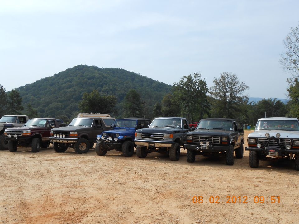 Several lifted Ford trucks and SUVs parked on dirt, with a wooded hill in the background