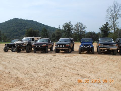 2013 Ford Bronco - A lineup of modified off-road vehicles, including a dark blue 1992 Ford Bronco, are parked in a dirt lot with a wooded hillside in the back…