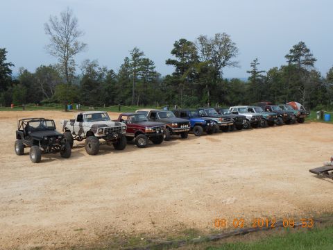 2013 Ford Bronco - A collection of modified off-road vehicles are lined up on a dirt lot, with trees and a cloudy sky in the background.