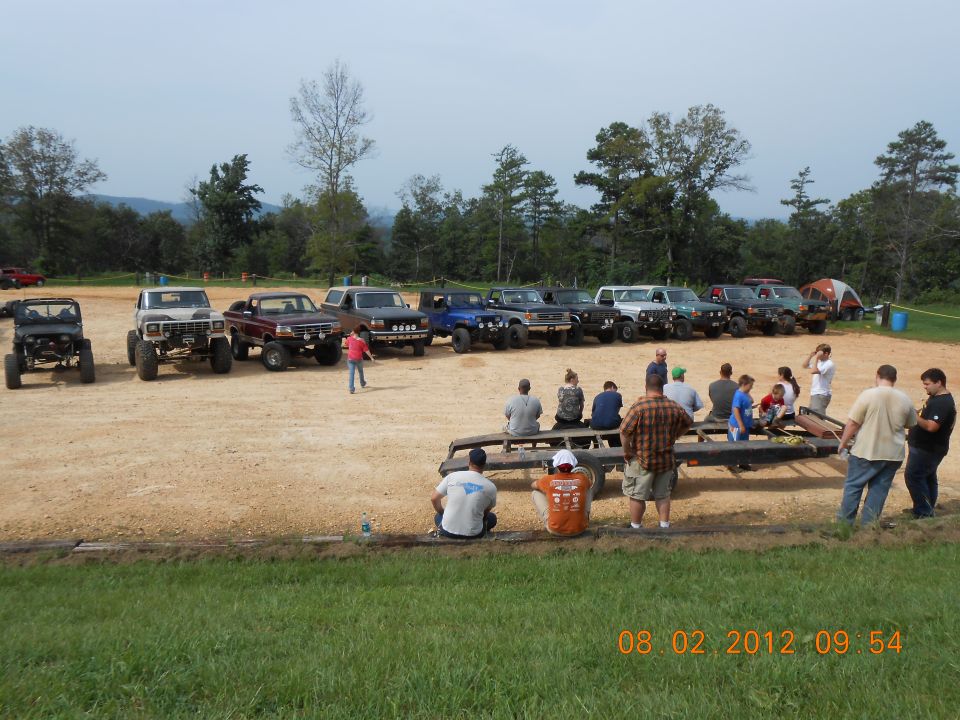 Several off-road trucks and SUVs parked in a dirt lot with people gathered around