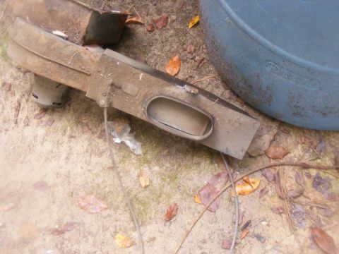 1978 Ford Bronco - A weathered car part, possibly a bumper or trim piece, lies on the ground next to a blue barrel.