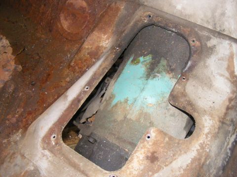 1978 Ford Bronco - Close-up view of a heavily rusted metal panel with a cutout revealing a teal-colored component and some wiring underneath.