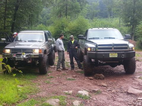 1987 Toyota 4Runner - Two lifted trucks, a Toyota 4Runner and a Dodge Ram, are parked on a dirt path in a wooded area.
