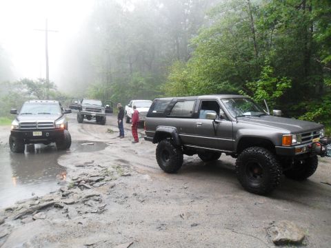1987 Toyota 4Runner - Several off-road vehicles are gathered on a wet, muddy path surrounded by trees on a foggy day.